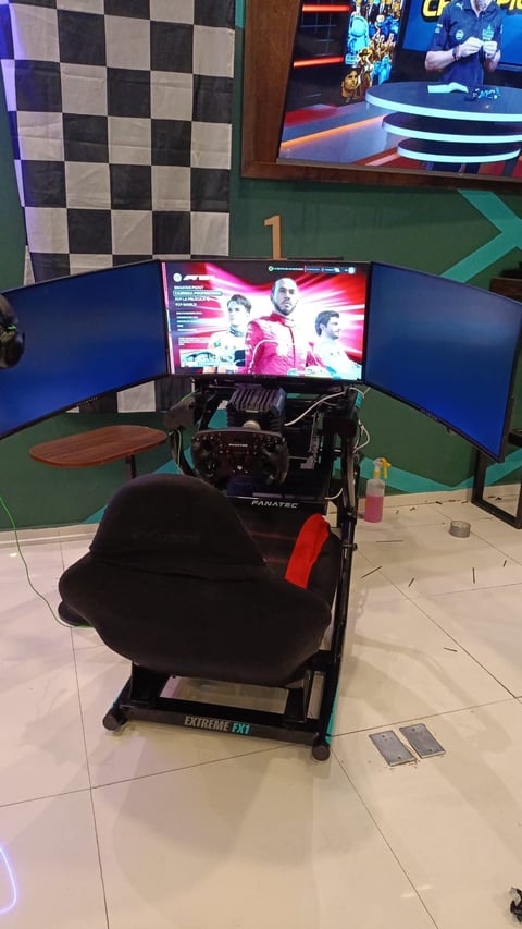 Gaming setup with triple blue monitors displaying racing game, Fanatec racing wheel, black racing seat, in gaming lounge with checkered wall backdrop