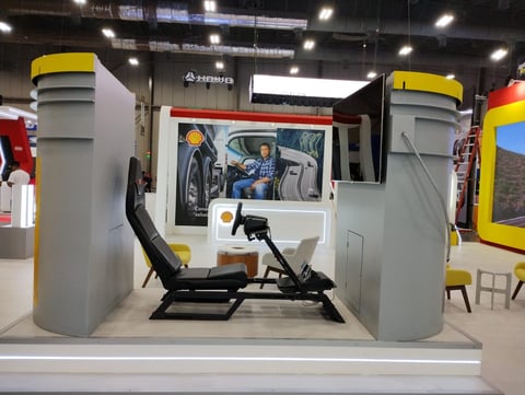 Racing simulator cockpit with steering wheel and pedals positioned between two gray pillars with yellow tops at an indoor exhibition
