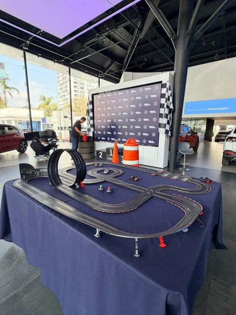 Slot car racing track display on blue table at automotive event with sponsor backdrop and showroom in background